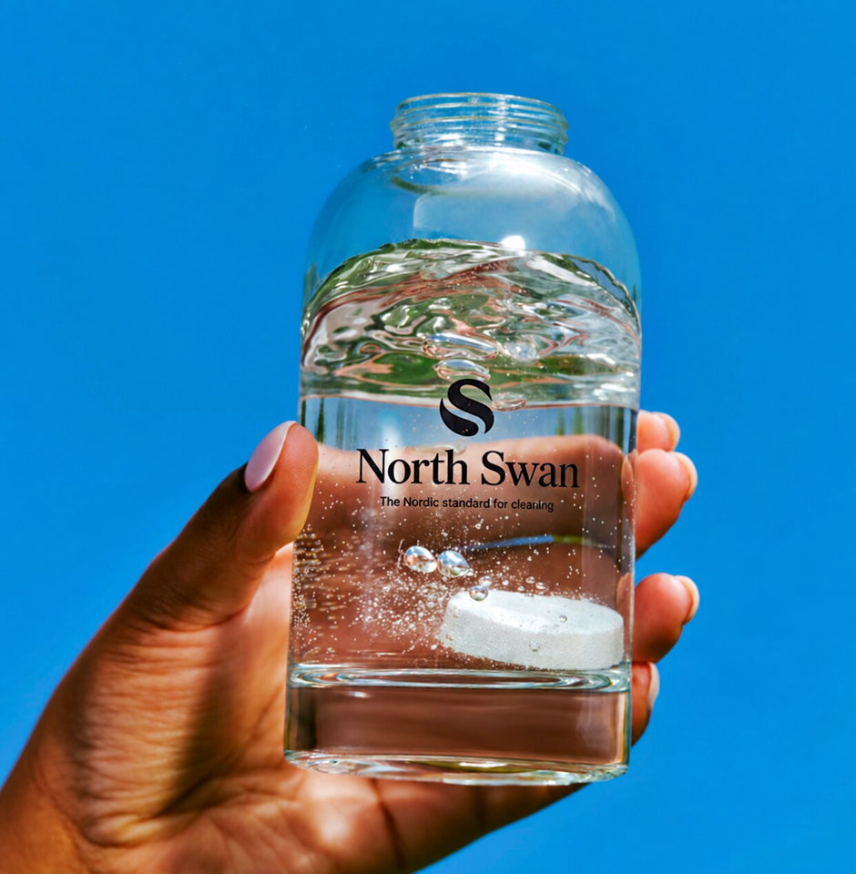 Hånd holder North Swan glasflaske med en sæbetablet, der opløses, op mod en blå himmel